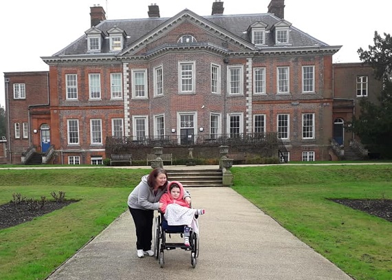 Chloe: young girl in a wheelchair with adult, in front of Mansion building at The Children's Trust