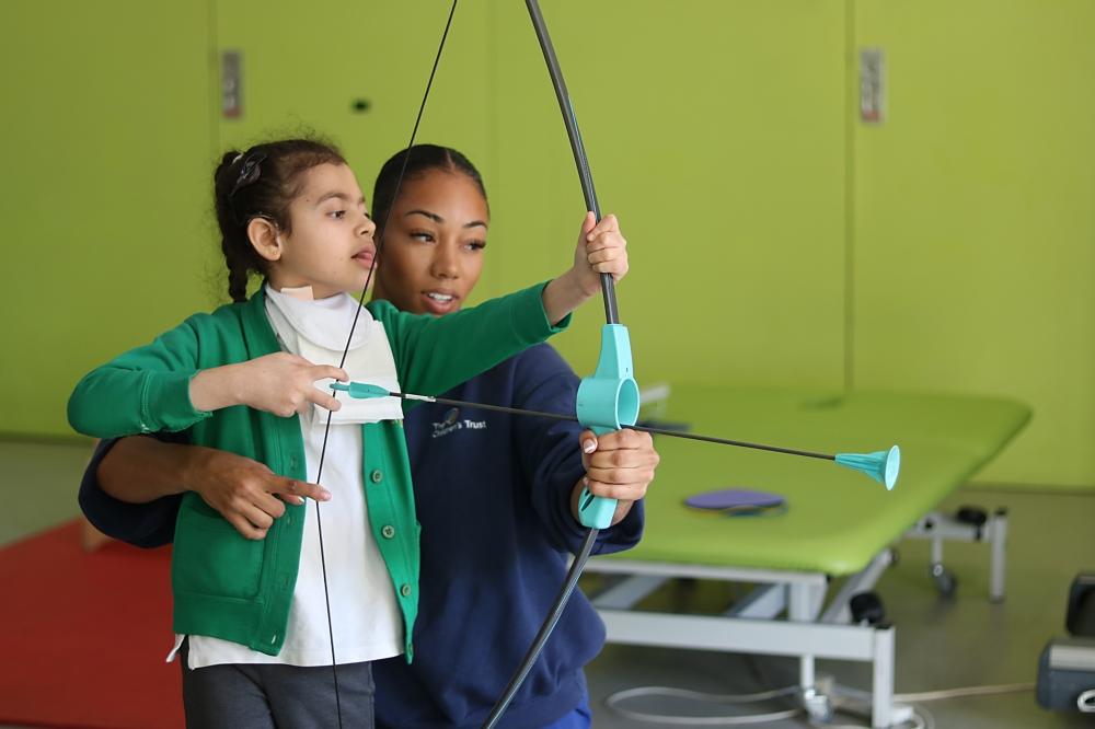 Shakeerah aiming with a bow and arrow game, supported by her physiotherapist