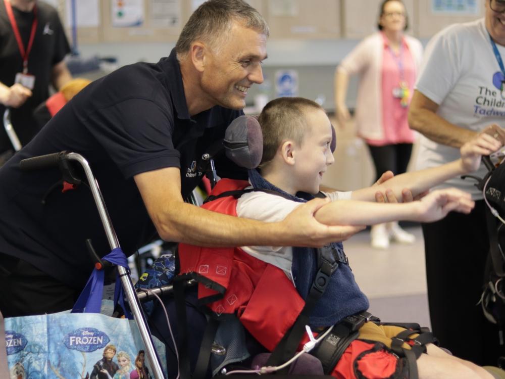 A boy in a wheelchair stretching his arms and smiling. His Dad is holding his arms and smiling.