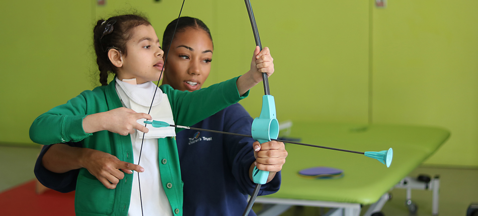 Shakeerah aiming with a bow and arrow game, supported by her physiotherapist