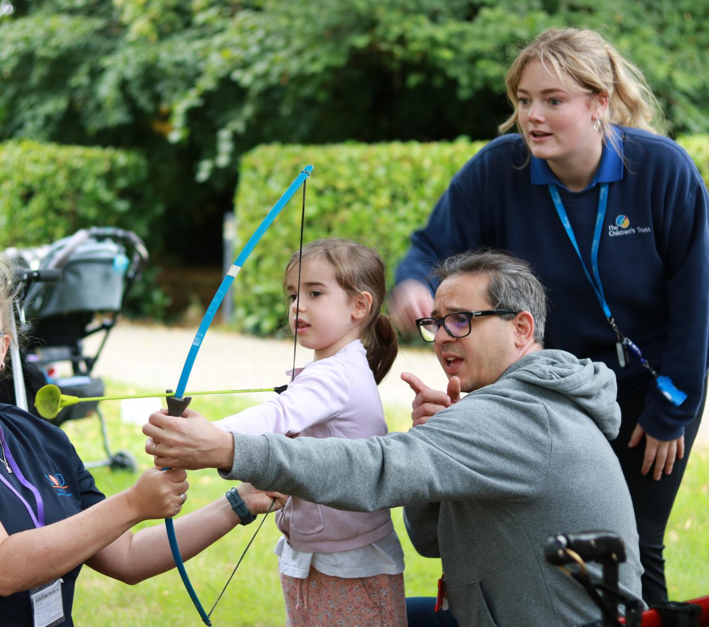 Child holding a bow and arrow with her dad pointing behind her 