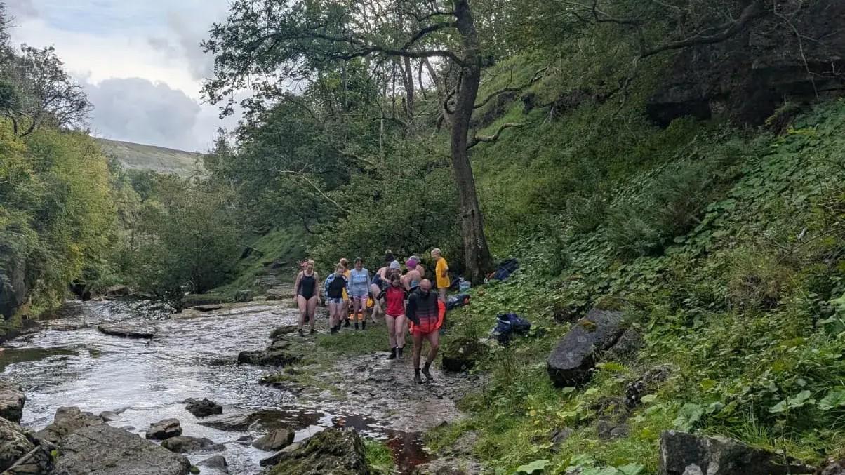 hikers along the side of a river running through a lush green mountainous valley