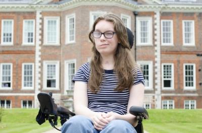 A smiling teenage girl in a wheelchair sits in front of an old manor house
