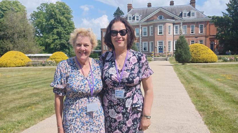 Two ladies standing side by side smiling at the camera, in the background is a mansion house