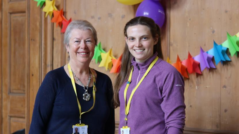 Two volunteers smiling at the camera, there are balloons and a party banner behind them