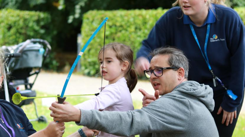 Child holding a bow and arrow with her dad pointing behind her 