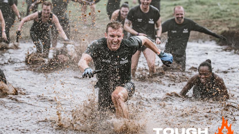 smiling man splashing through a muddy pool of water with fellow racers falling behind him