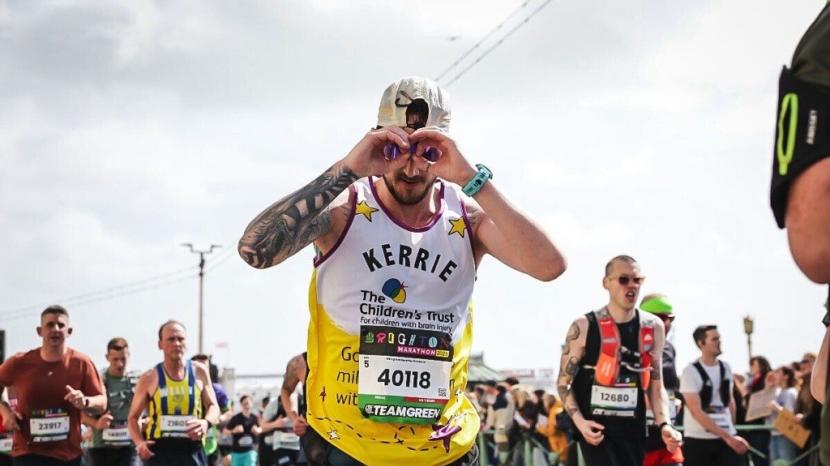 Runner with sleeve tattoo in The Children's Trust yellow and white running vest, with a green watch and cream baseball cap, other men running in the background against a grey sky