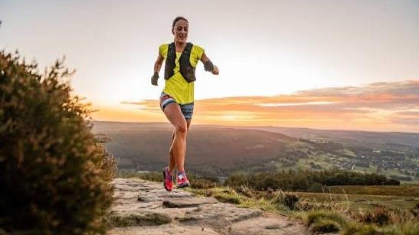 Female runner in yellow top, stripey shorts and pink trainers at sunset running along a hillside path flanked with lush green grass and a picturesque orange sky